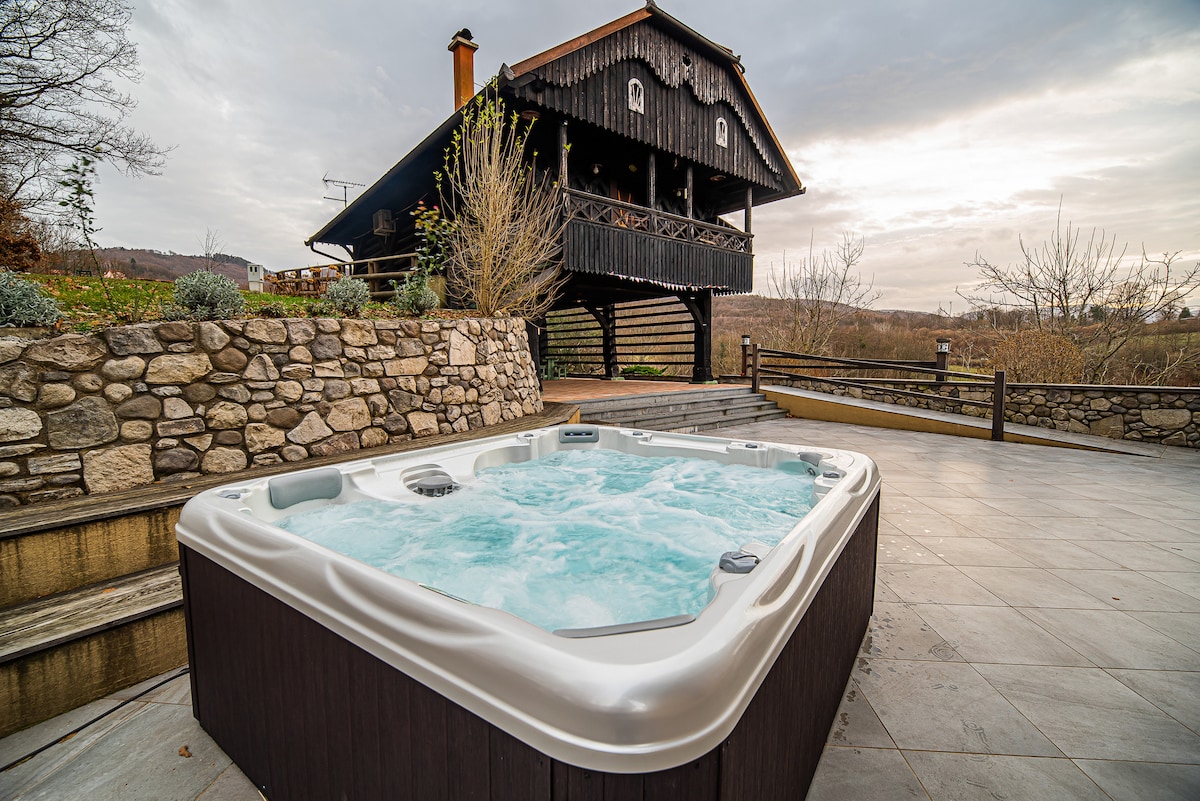An outdoor hot tub is positioned on a spacious terrace with stone and tiled flooring. The rustic wooden house, consisting of dark timber, stands prominently in the background, surrounded by nature. Scenic views of the landscape are visible beyond the terrace.