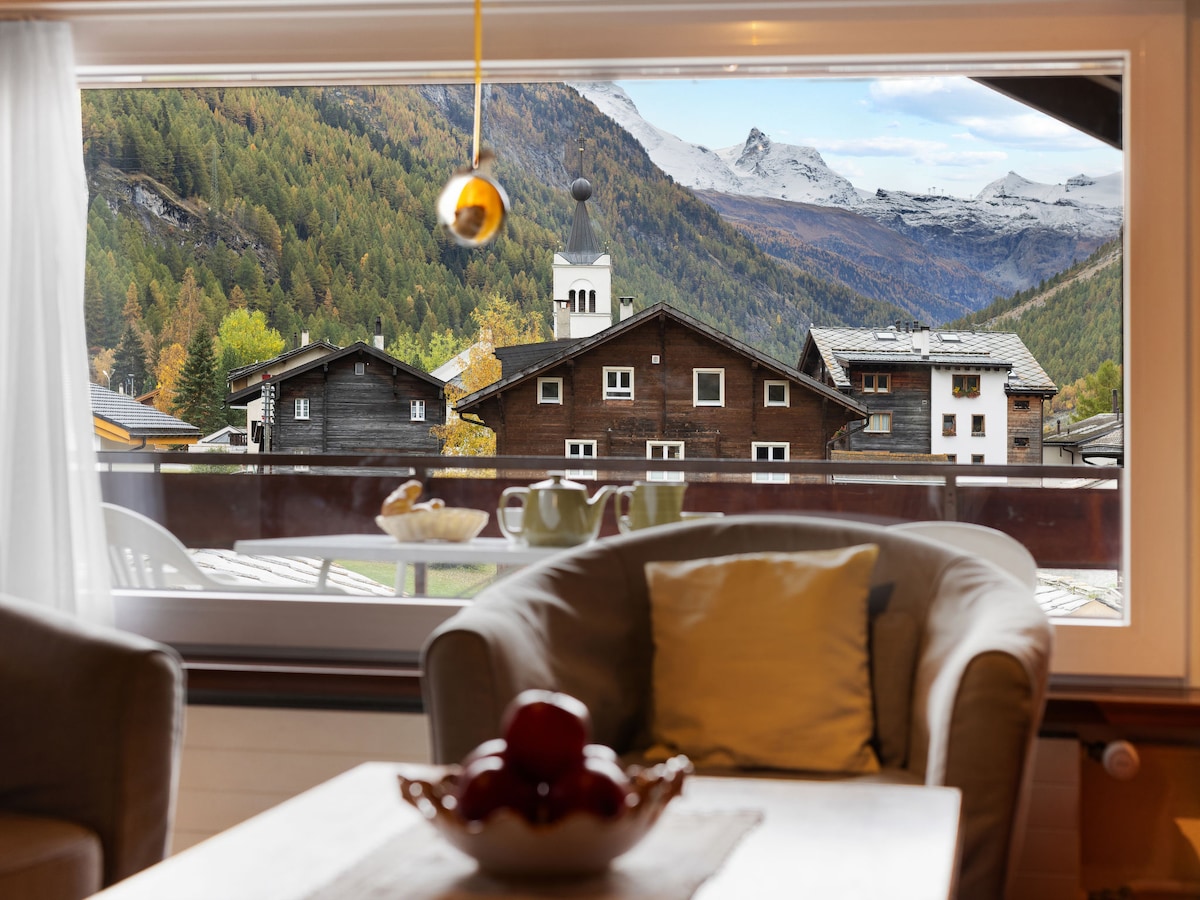 A view is captured from the window, revealing charming wooden buildings set against a mountainous backdrop. Fall foliage blankets the landscape in warm hues. A small table is visible, holding a bowl of red apples, alongside a coffee set, enhancing the inviting atmosphere.