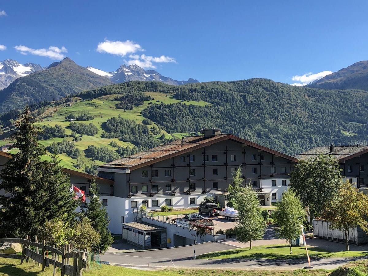 The image captures a scenic view of the surrounding landscape, featuring the apartment building in the foreground. Lush green hills and towering mountains are visible in the background under a clear blue sky, creating a serene atmosphere.