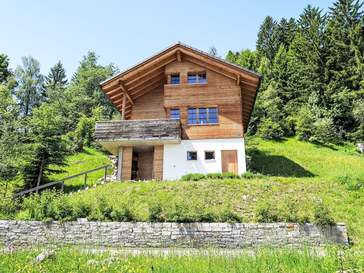 The chalet showcases a wooden exterior with a sloping roof and large windows, set against a backdrop of lush greenery and pine trees. A small covered entrance is visible, leading up to the building, which is surrounded by meadows.