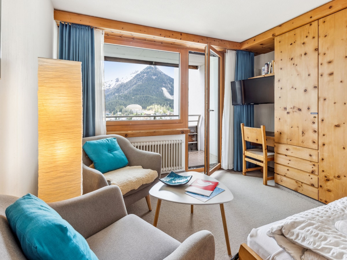 The living area features rustic furnishings with a comfortable chair and sofa, a round coffee table, and a flat-screen TV mounted on a wooden cabinet. Natural light fills the space through a large window and balcony door, offering views of the mountains.