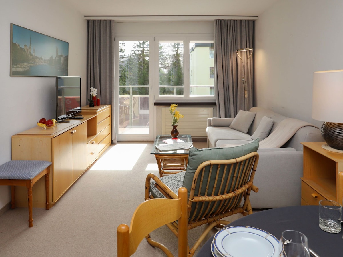 A living and dining area features a light-colored sofa and armchair, accompanied by a wooden dining table. Natural light fills the space through large windows, leading to a balcony. A TV is positioned on a wooden console, enhancing the room's inviting atmosphere.