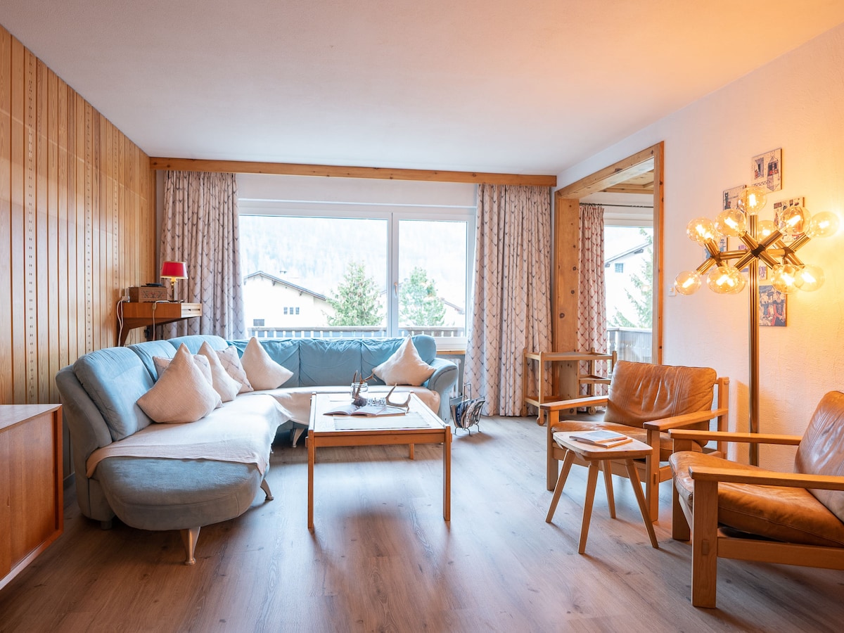A spacious living area features a combination of soft blue and wooden furnishings. Large windows provide natural light and a view of the surrounding mountains. A coffee table sits at the center, complemented by comfortable seating arrangements, including armchairs and a cozy sofa.
