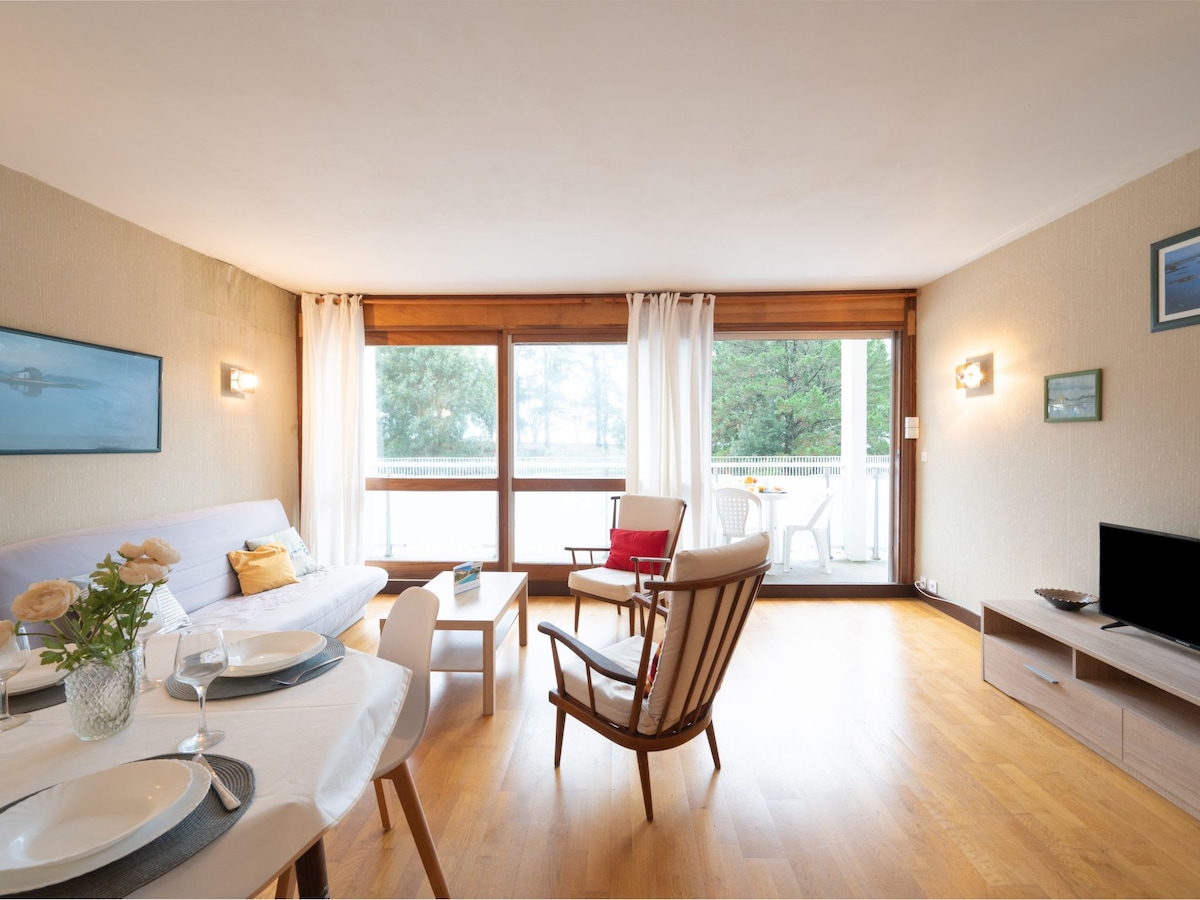 The living area is characterized by practical furnishings, featuring a comfortable sofa and two armchairs arranged around a small coffee table. Large sliding doors allow natural light to fill the space, leading to a balcony with views of the surrounding trees.