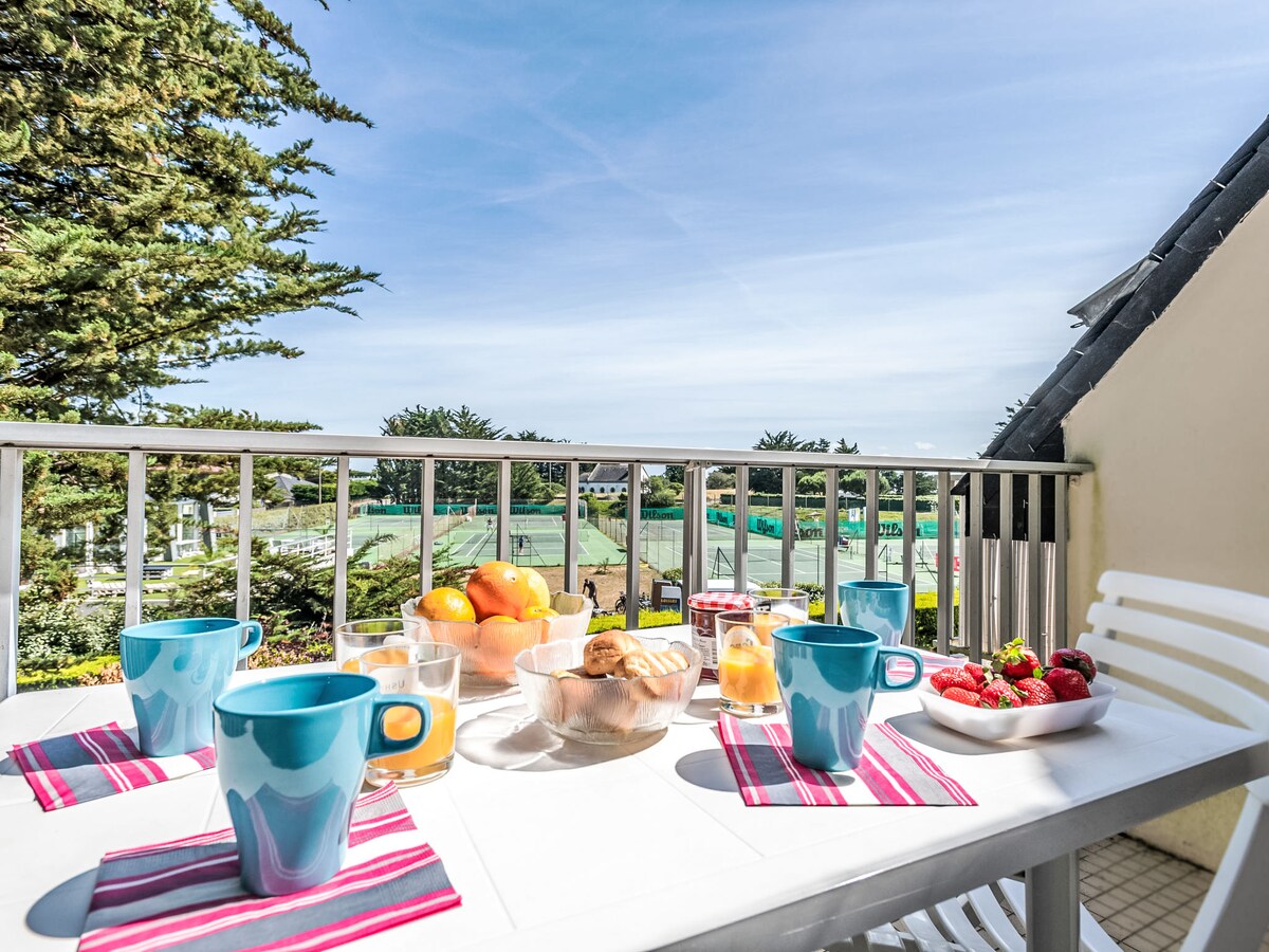 A dining table set outdoors is adorned with colored mugs, plates of fresh fruit, and baked goods. The balcony offers a view of green courts and trees beyond, creating a pleasant environment for meals and relaxation.