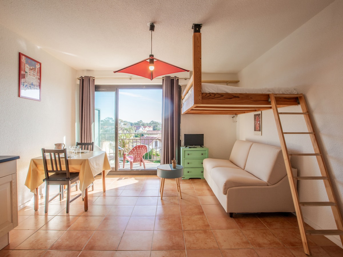 A living area features a white sofa, a round table with chairs, and a kitchenette along one wall. Above, a lofted bed accessed by a ladder creates an open sleeping space. Floor-to-ceiling glass doors lead to a small balcony with a view.