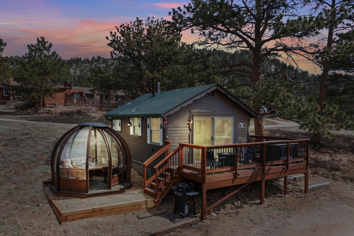 A cozy cabin is set amidst a serene landscape, featuring a unique heated dining globe on a deck. The cabin's welcoming exterior and large windows evoke a sense of comfort, as trees surround the property, offering privacy and a natural feel.
