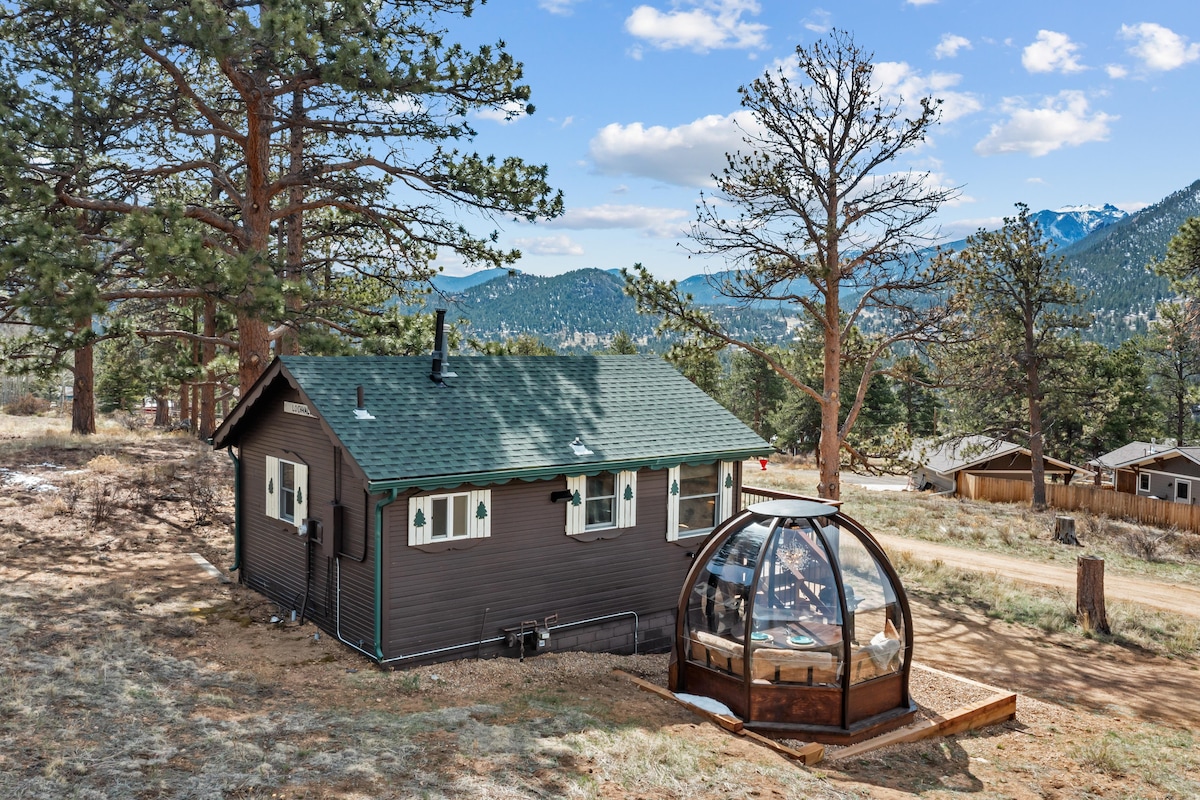 A charming cabin with a brown exterior is nestled among trees, showcasing a unique heated dining globe nearby. The backdrop features expansive mountain views under a clear blue sky, while the landscape is dotted with natural vegetation and distant structures.