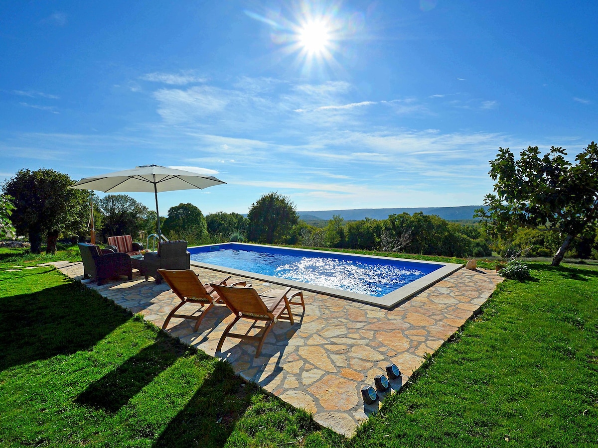 The image depicts a sunny outdoor area featuring a rectangular swimming pool surrounded by natural stone tiles. Two deck chairs are positioned beside the pool under the shade of a large umbrella. Lush green grass and trees enhance the tranquil setting, with a clear sky above.