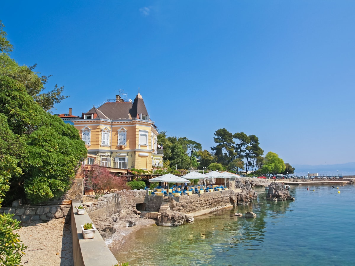A historic apartment house with yellow exterior is situated along the coastline. The building features large windows and a distinctive roof. Adjacent, there are outdoor seating areas set among lush greenery. Calm waters reflect the clear blue sky, enhancing the serene seaside environment.