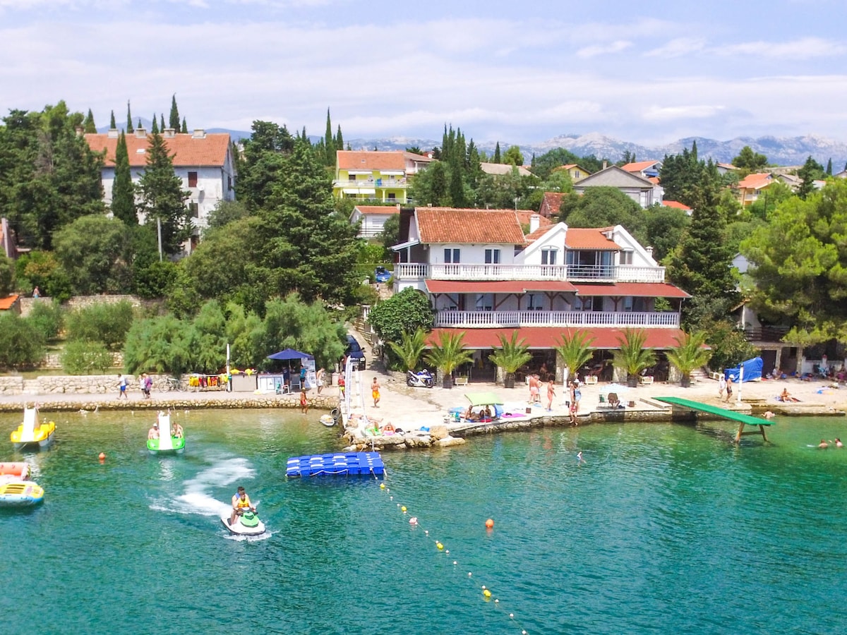 The image captures a seaside scene featuring a vibrant waterfront and a cozy apartment building. Clear turquoise waters are visible, with various water activities taking place. The surrounding area boasts lush greenery and well-maintained pathways, creating an inviting atmosphere by the beach.