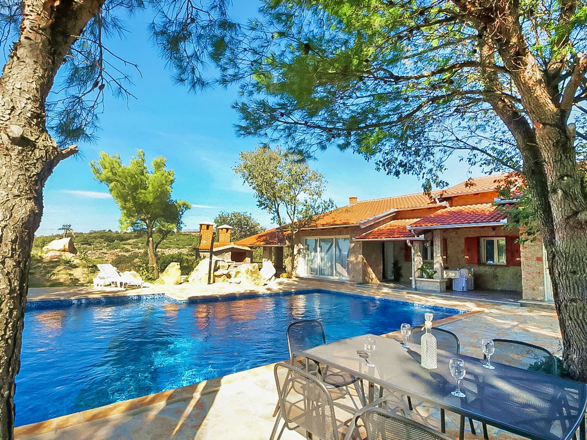 A spacious outdoor area is showcased, featuring a large swimming pool with a clear blue surface. Sun loungers are positioned nearby under the shade of trees, and a dining table with several chairs is visible, providing a comfortable space for outdoor meals.