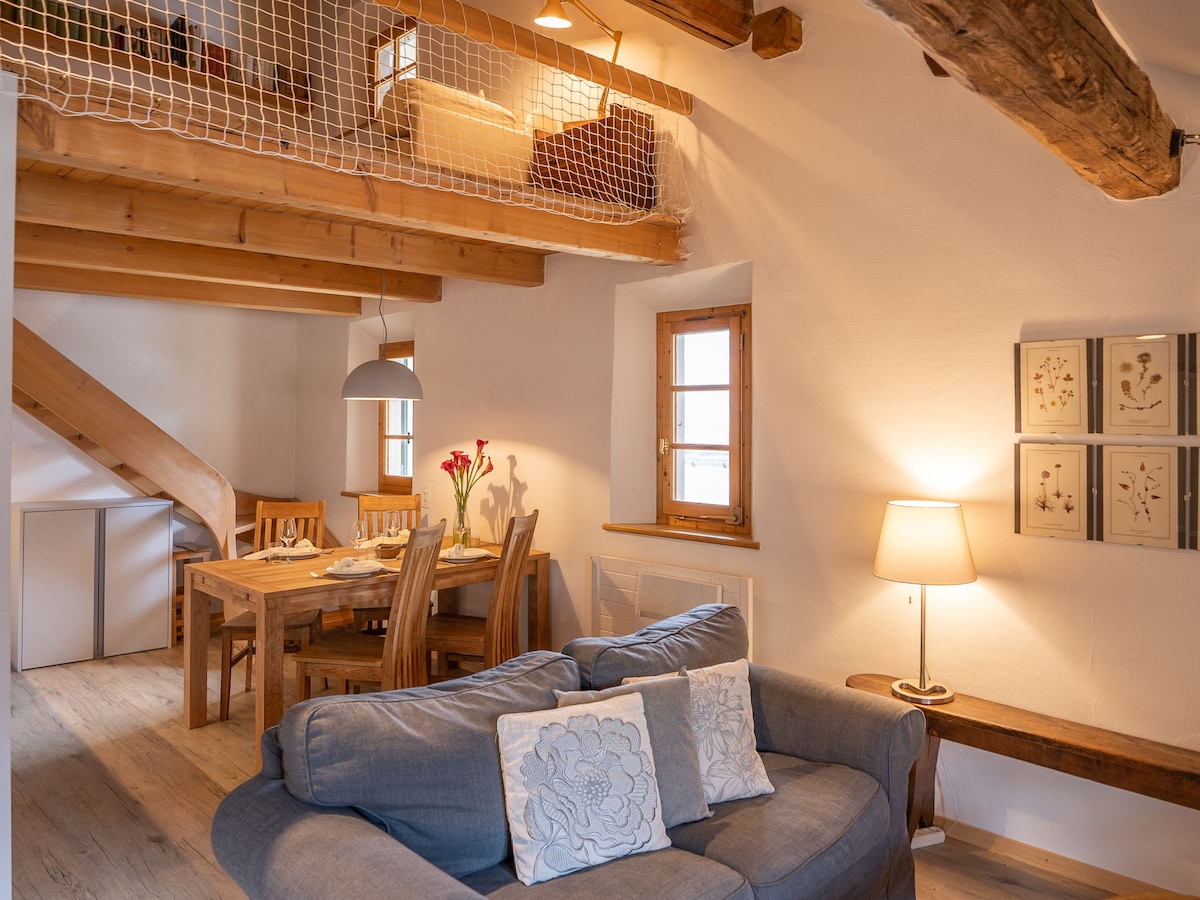 The living area displays a cozy seating arrangement with a blue sofa adorned with decorative pillows. A dining table with wooden chairs is seen, illuminated by overhead lighting. Stairs lead to an upper level, where a gallery is visible along the wall.