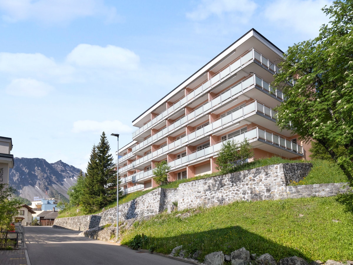 A three-story apartment block is set against a backdrop of mountains. The building features multiple balconies with glass railings and light-colored walls. A stone wall borders the property, while greenery and trees enhance the outdoor area.