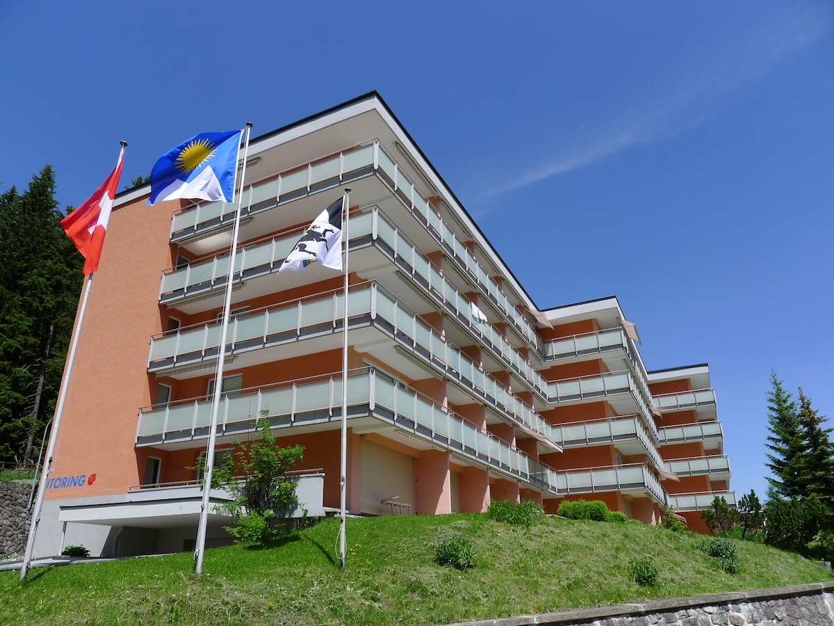 The apartment block features a three-story building with a light orange facade and multiple balconies. Flags from Switzerland and the local resort flutter in the foreground. Lush greenery surrounds the base, contributing to the tranquil setting amidst a clear blue sky.