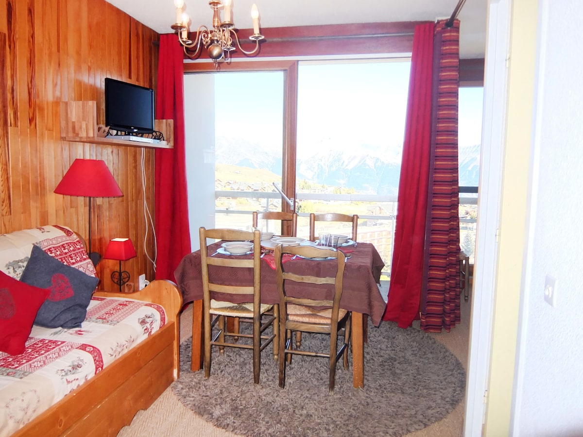 A welcoming living and dining area features a wooden wall and large panoramic window showcasing mountain views. A pull-out bed is positioned next to a red lamp, and a dining table is set for four. Rich red curtains frame the window, enhancing the cozy ambiance.