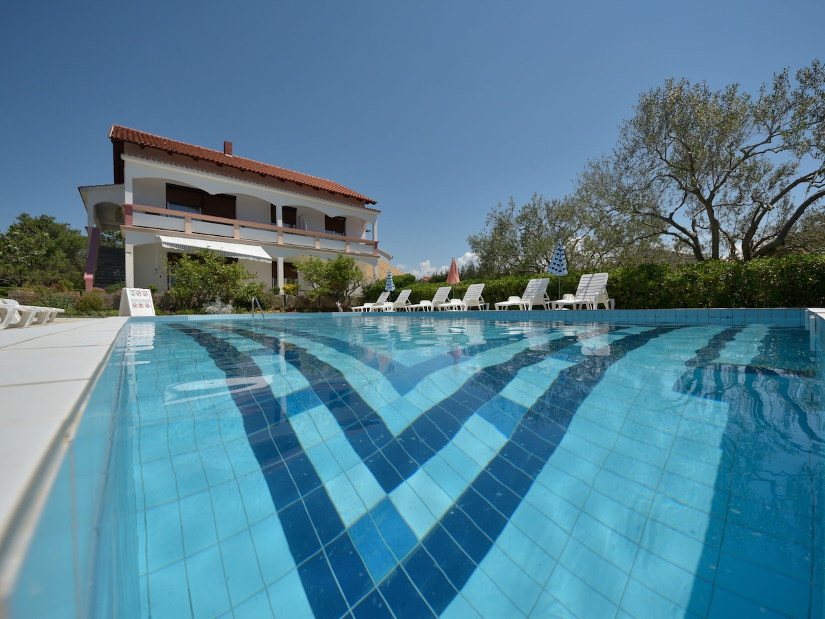 A swimming pool features clear blue water with a tiled floor reflecting light. Sun loungers line the poolside, situated against a backdrop of a two-story house with a red-tiled roof and surrounded by greenery.