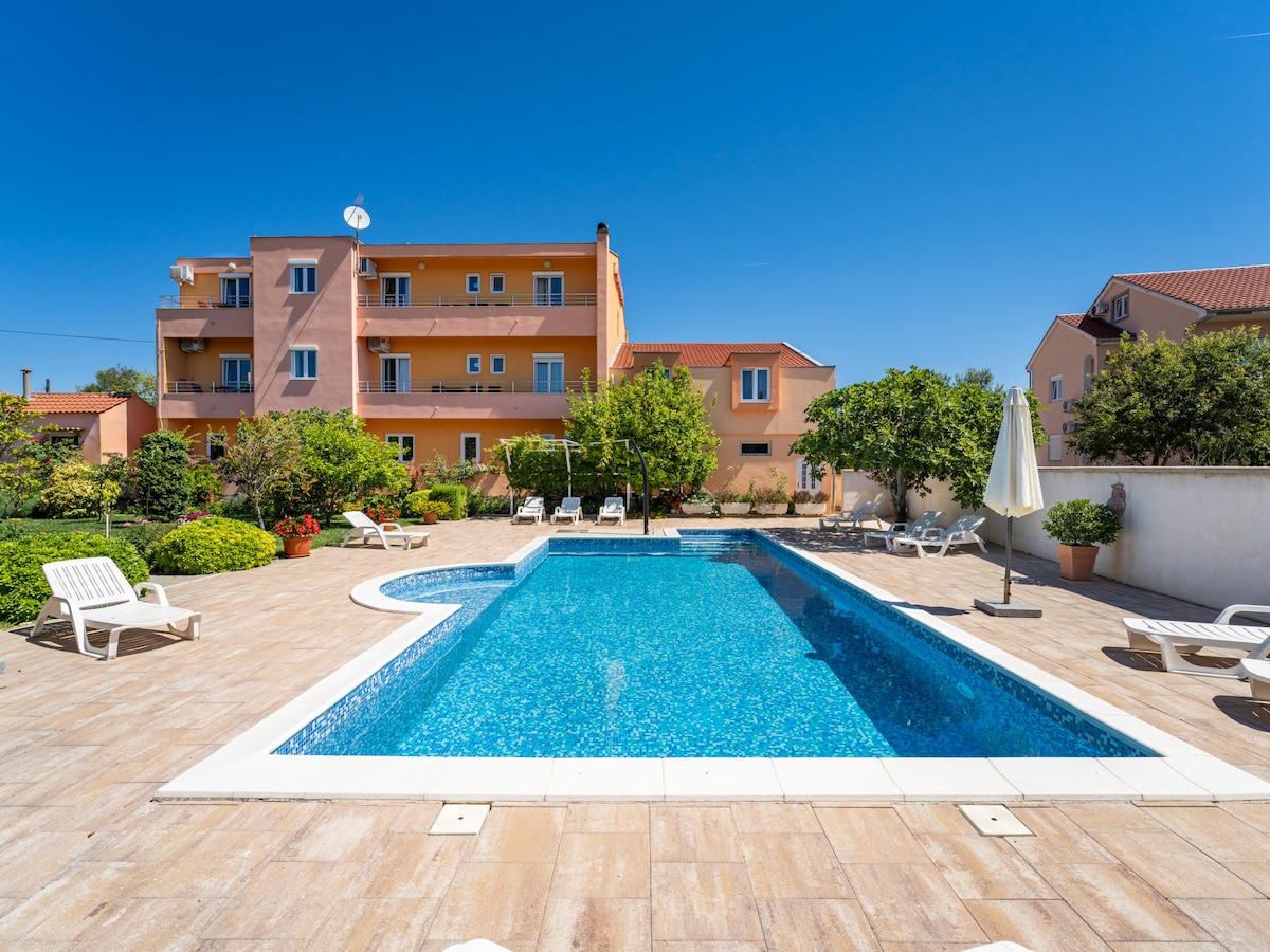A clear blue swimming pool is surrounded by a spacious patio adorned with lounge chairs and greenery. The two-story apartment building is visible in the background, framed by vibrant plants and blooming flowers. Sunlight reflects off the water, creating a bright ambiance.