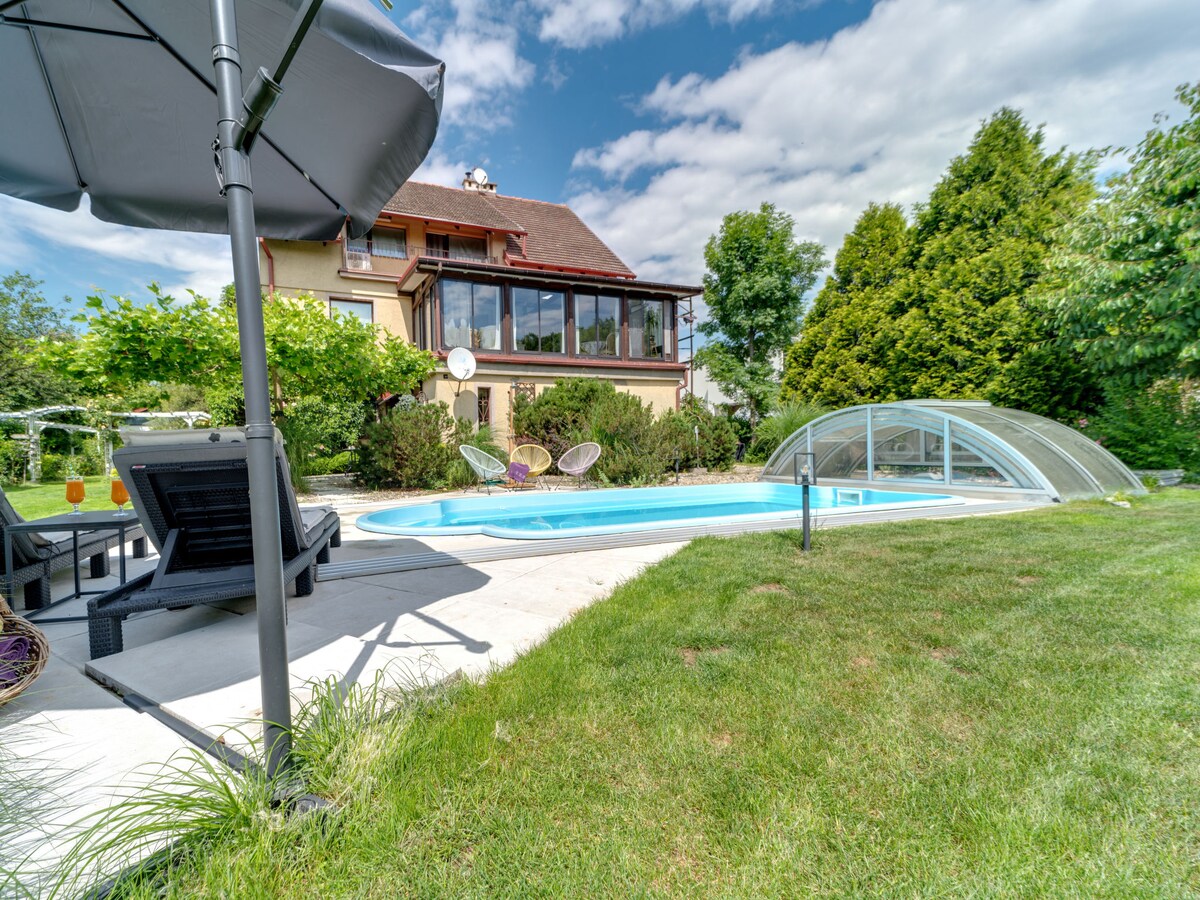 A fenced garden features an angular heated swimming pool, surrounded by neatly maintained grass. A shaded area with seating is visible, complemented by a light canopy. The background showcases a two-story house with a large glass-enclosed terrace, set against a clear sky and greenery.