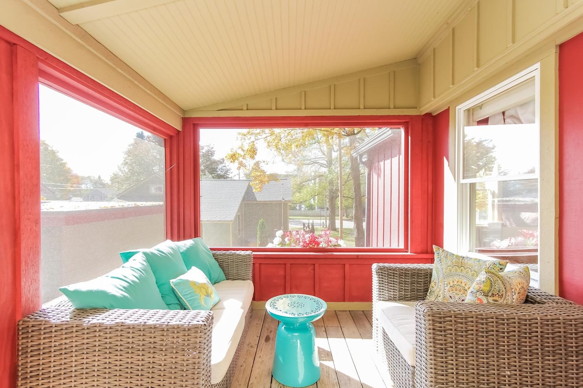 A screened-in porch features comfortable wicker seating with colorful cushions, alongside a small turquoise table. Large windows allow natural light to brighten the space, while views of the surrounding area can be seen through the screens.