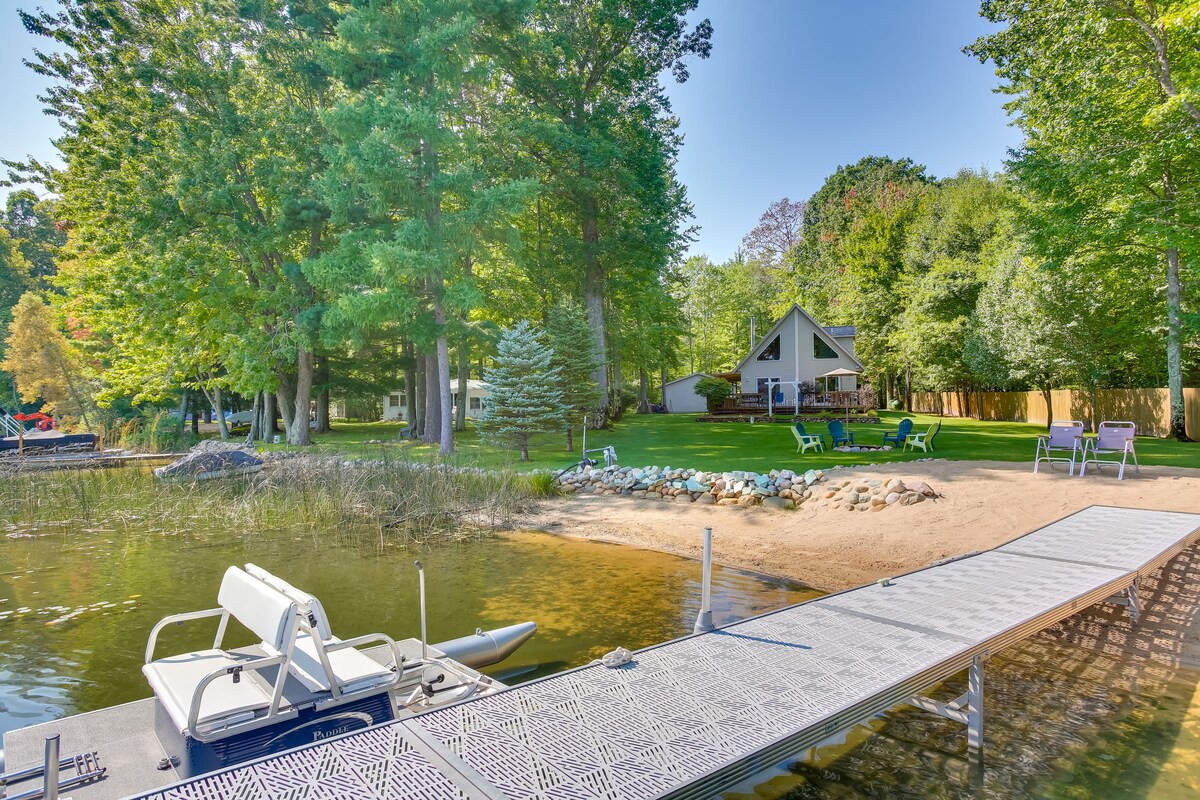 A view from the dock showcases the lakefront property surrounded by lush greenery. A sandy area is visible near the water, along with colorful seating options scattered across a spacious lawn. The serene lake reflects the sky, providing a tranquil atmosphere.
