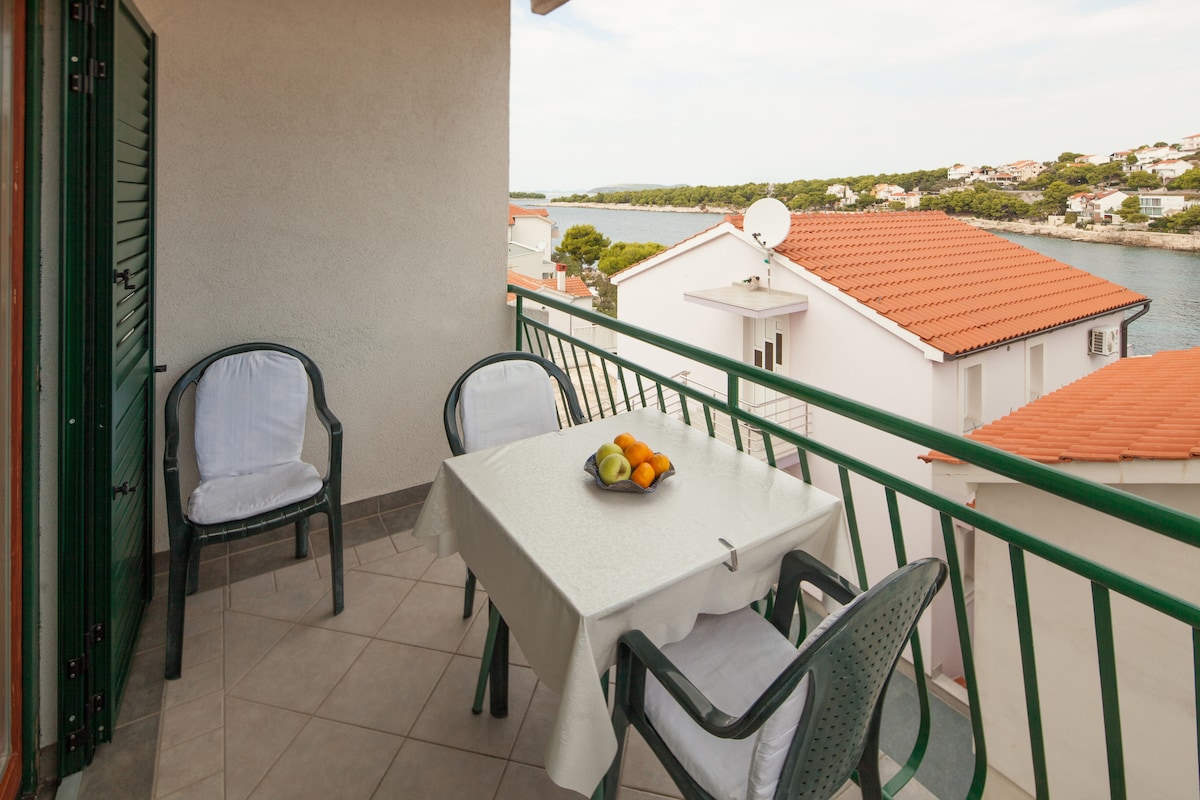 A private terrace features a dining table with a fruit centerpiece. Two chairs with cushions are positioned around the table, and a view of the sea and surrounding houses is visible in the background.