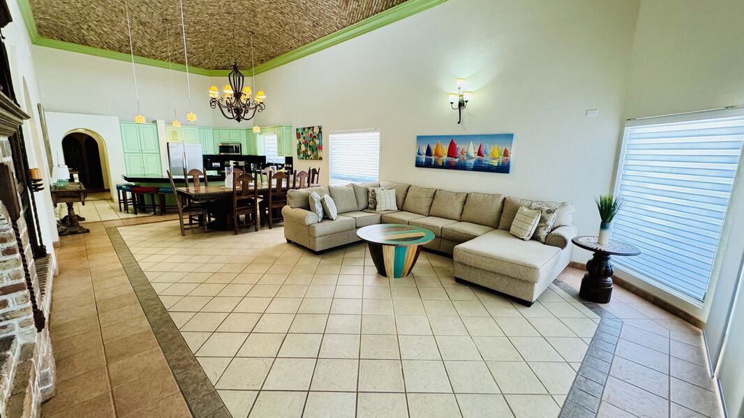 An open living area showcases a large sectional sofa and a coffee table with vibrant colors. Light-colored walls are complemented by a chandelier and artwork featuring sailboats. A dining table occupies the space behind, with ample seating for guests, and natural light enters through large windows.