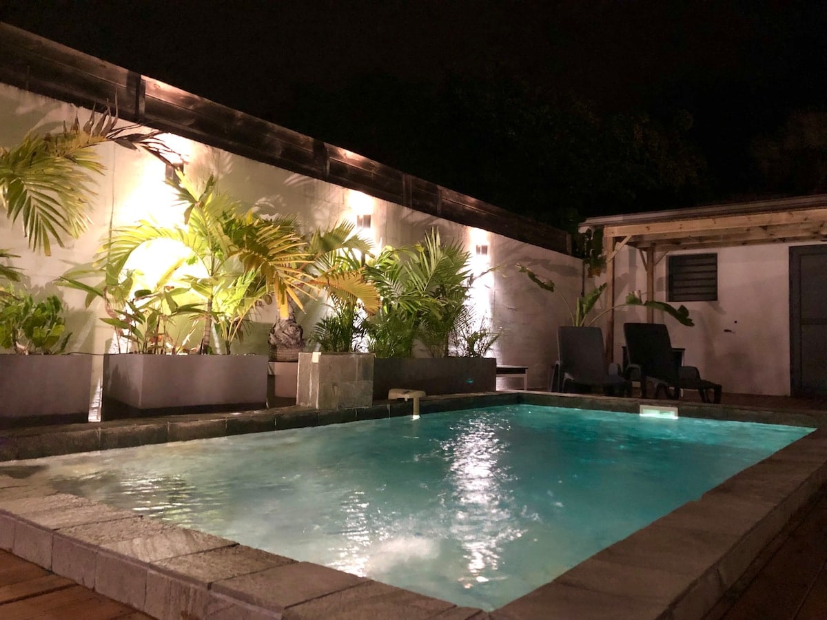 A tranquil common pool is illuminated at night by soft lighting, surrounded by lush greenery in planters. Comfortable lounge chairs are positioned nearby, inviting guests to relax in a serene environment.