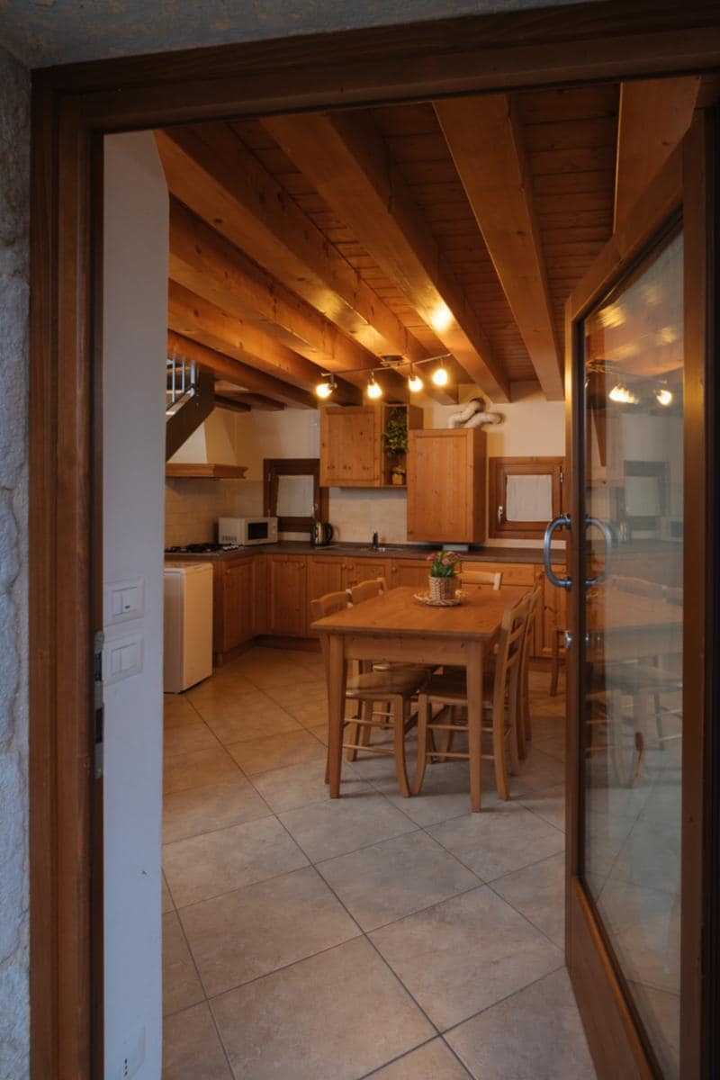 A warm kitchen area is visible through a wooden door frame. Light wooden cabinetry complements the stone walls, and a dining table surrounded by chairs sits center stage, illuminated by overhead lights. Kitchen appliances and utensils are neatly arranged along the counters.