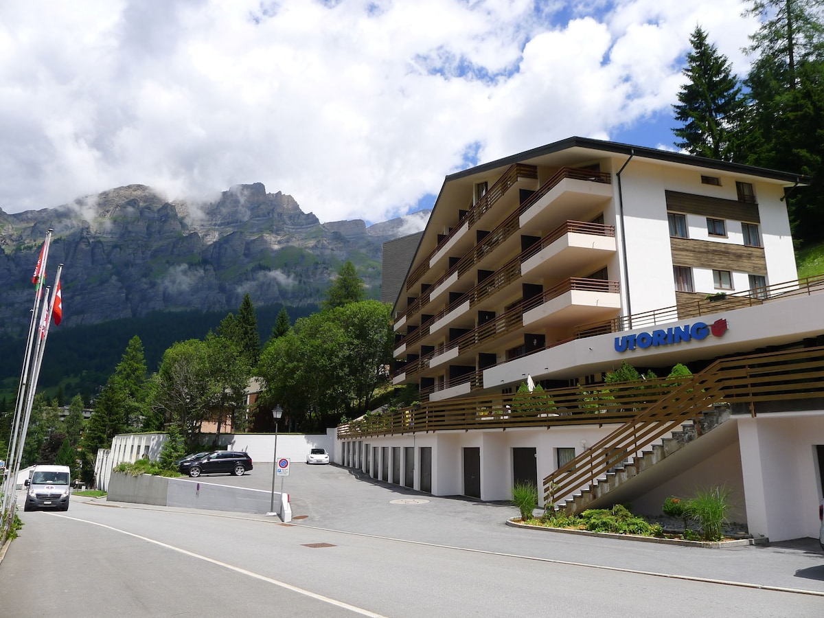 The apartment building is situated against a backdrop of towering mountains, featuring a modern architectural style with multiple balconies. Trees are visible around the property, and a spacious parking area can be seen in the foreground, alongside a road leading to the entrance.
