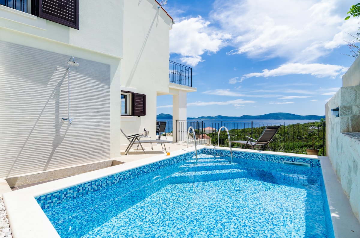 A clear blue swimming pool is framed by a sunlit terrace, featuring two lounge chairs positioned for relaxation. The pool area is complemented by sweeping views of the landscape and distant sea, with a light-colored wall and window providing a bright backdrop.