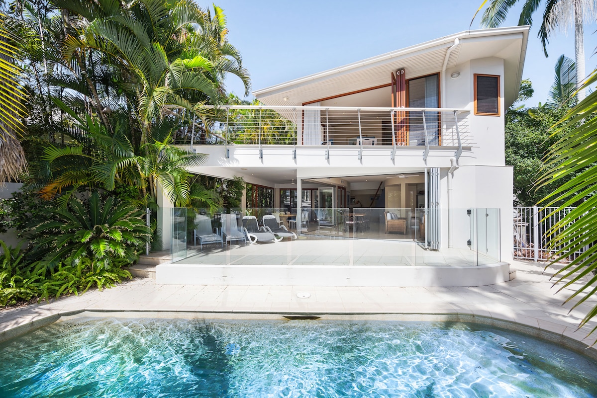 A modern multi-level home with a spacious terrace overlooking a sparkling pool is surrounded by lush tropical landscaping. The glass balustrade enhances views of the outdoor area, which includes sun loungers. Large glass doors on the upper level allow ample natural light into the interior spaces.