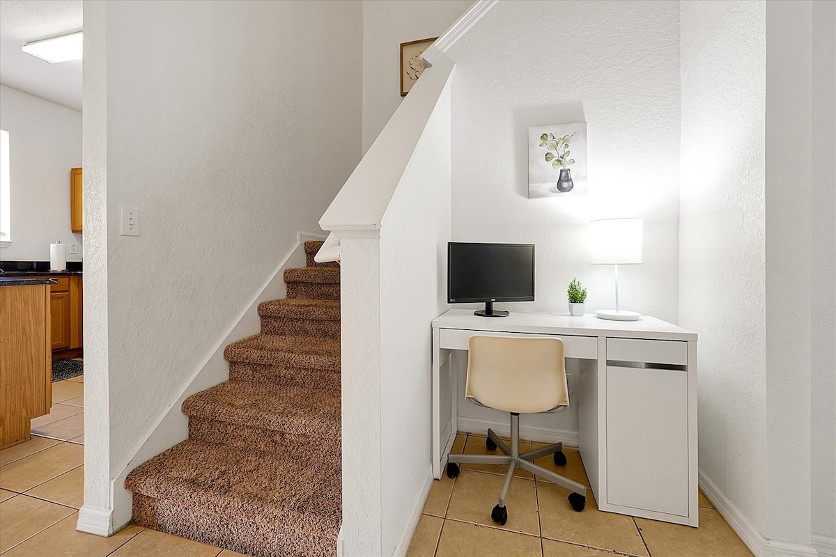 A compact workspace is positioned conveniently by the staircase, featuring a small desk with a chair and a monitor. Soft light from a lamp casts a warm glow, highlighting the clean white walls and decorative plant. The flooring transitions from tiles to carpeted stairs.