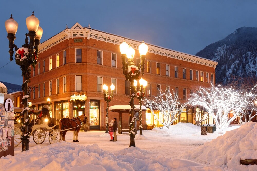 A historic brick hotel is set against a snowy landscape in the evening. Streetlamps adorned with festive decorations illuminate the scene, while a horse-drawn carriage is stationed nearby. Snow-covered trees glisten, and the structure's warm lights create a welcoming ambiance.