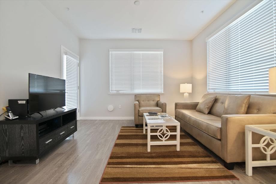 A modern living space features a plush sofa paired with a stylish coffee table. Natural light streams through large windows adorned with blinds, and a flat-screen TV is positioned on a sleek console against the wall. Soft neutral tones create a serene atmosphere.