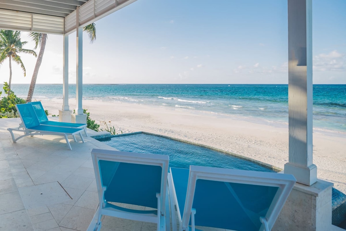 Two lounge chairs with bright blue cushions are positioned on a deck overlooking the ocean. The infinity pool seamlessly merges with the horizon, where the waves gently lap against the shore. Palm trees sway softly in the background under a clear blue sky.