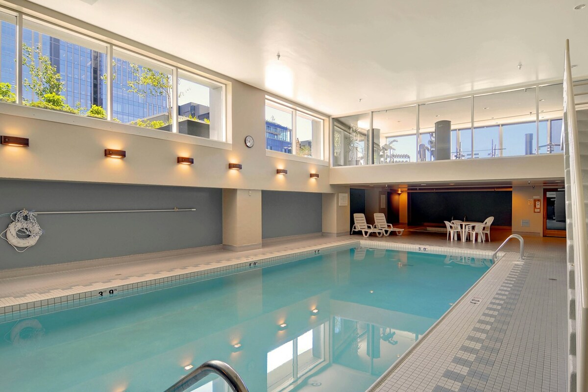 An indoor pool is featured with lap swimming lanes, surrounded by large windows that allow natural light to brighten the area. Relaxing seating is available nearby, complemented by gentle lighting on the walls, creating a serene atmosphere.