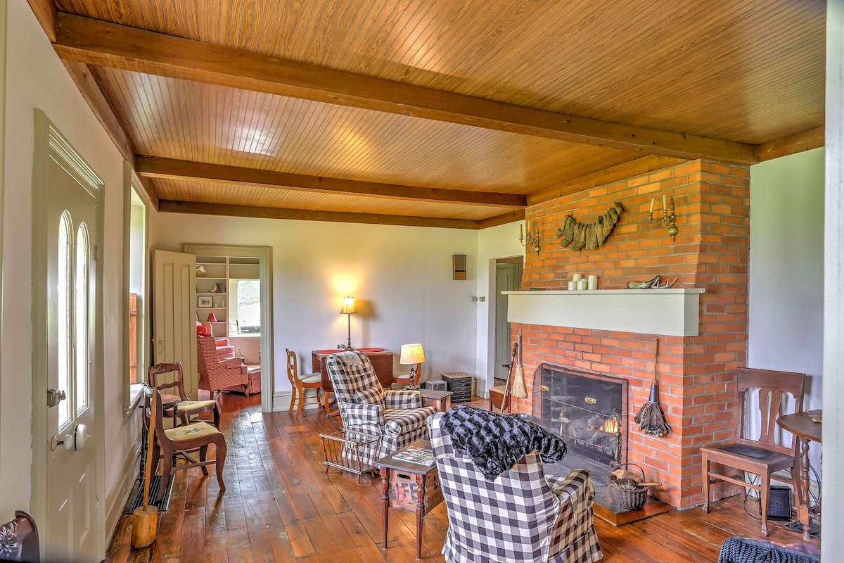 A warm living area features wooden beams overhead and a brick fireplace as a focal point. Comfortable seating arrangements include patterned armchairs and a classic rocking chair. Natural light fills the space through the nearby window, enhancing the room's inviting character.