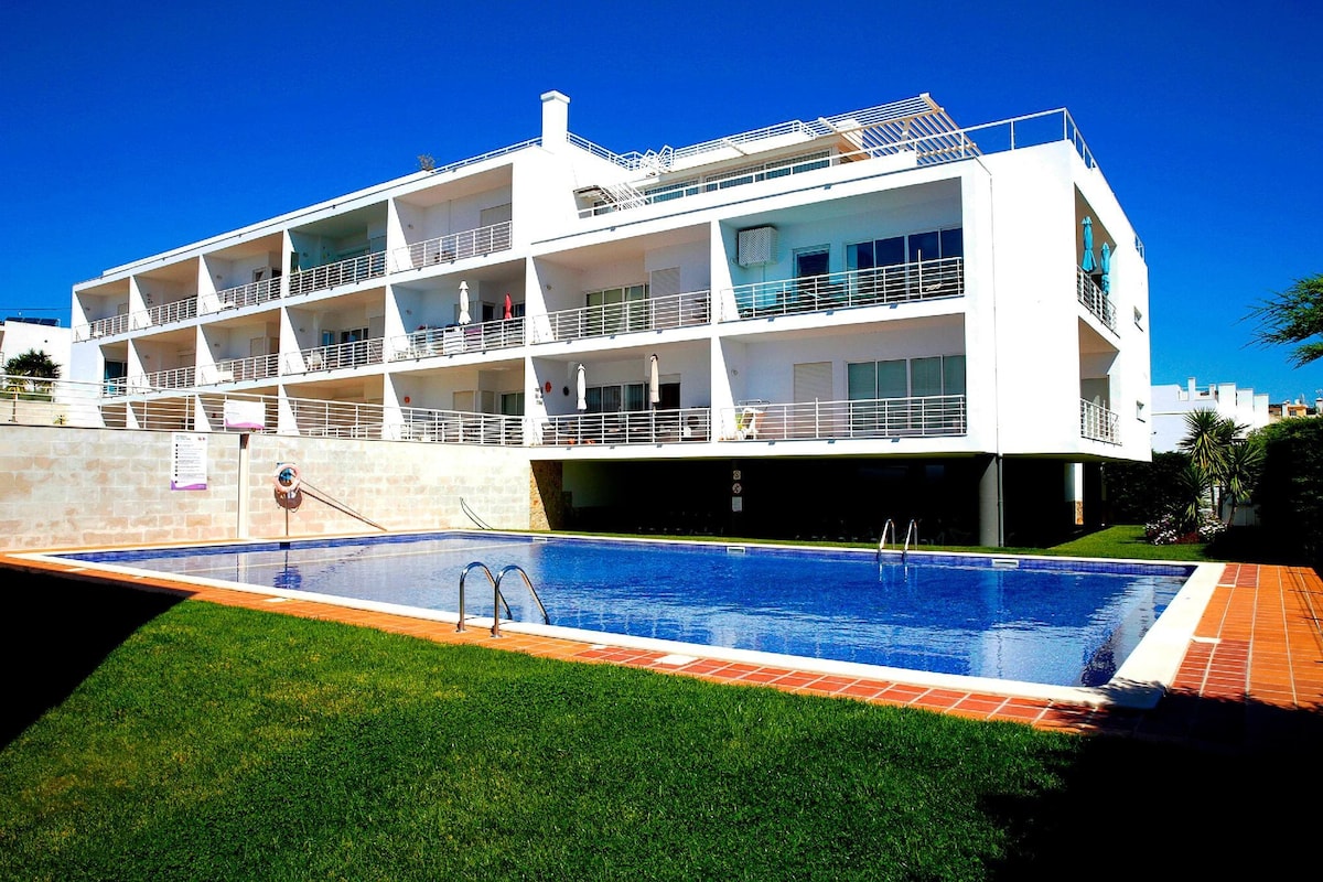 The modern condominium features a spacious swimming pool surrounded by lush green grass. The white-painted building with multiple balconies showcases large windows, providing a bright and open appearance. Sun loungers are placed on the poolside, inviting relaxation under clear blue skies.