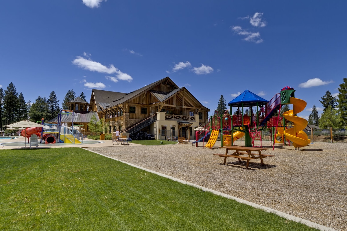 A spacious playground area is visible, featuring colorful slides, climbing structures, and swings. Nearby, a large wooden structure accommodates picnic tables. A well-maintained lawn surrounds the area, with verdant trees and clear blue skies enhancing the inviting outdoor atmosphere.