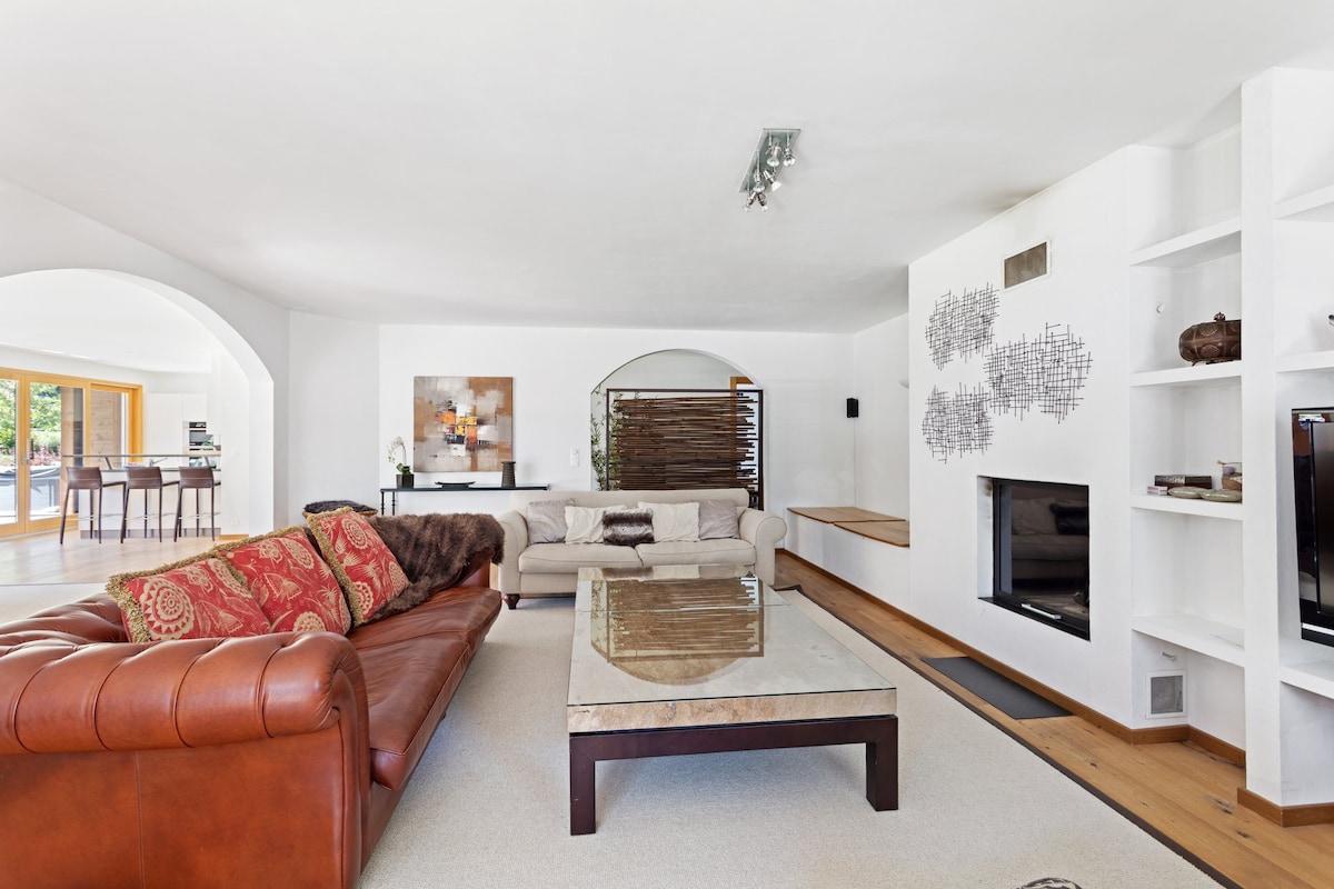 A spacious living room features a mix of a brown leather sofa and a light-colored fabric couch. A glass coffee table rests on a neutral rug, while the wall includes an entertainment unit with a television. Natural light floods the room through arched openings.