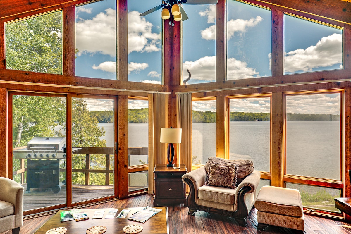 Large windows frame scenic lake views, allowing natural light to fill the open living space. Comfortable seating invites relaxation, while a decorative coffee table adds functionality. A gas grill is seen on the balcony, with trees complementing the serene outdoor surroundings.