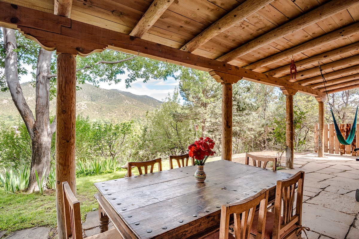 An outdoor dining area features a large wooden table surrounded by eight chairs, set beneath a wooden beam ceiling. A vibrant flower arrangement sits at the table's center. A hammock hangs nearby, with views of the hills and lush greenery in the background.