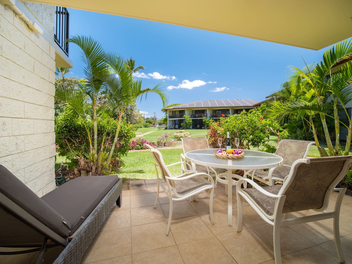 A shaded outdoor lanai features a round table surrounded by six chairs, offering a relaxed dining space. A reclining lounge chair is positioned nearby, with tropical greenery and colorful flowers framing the view. The setting provides a seamless indoor-outdoor experience.