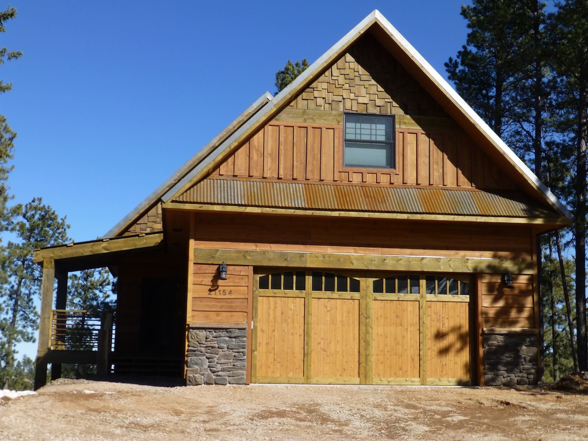 A charming cabin exterior features a combination of wood and stone elements. Double garage doors, framed by earthy tones, complement the gabled roof. The setting is surrounded by tall trees under a clear blue sky, suggesting a serene retreat in nature.