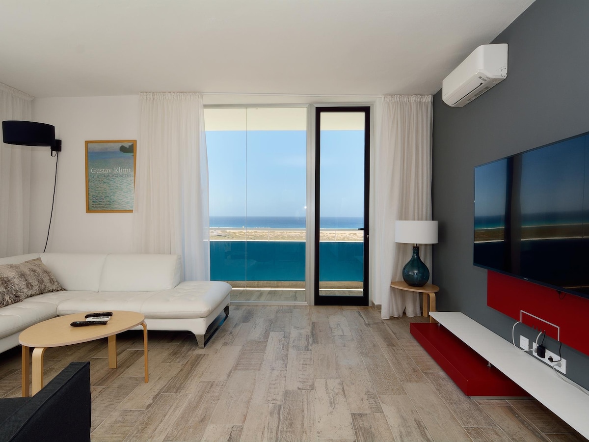 A bright living room is presented with a large sectional sofa and a side table. Floor-to-ceiling windows reveal expansive sea views, allowing natural light to fill the space. An entertainment system is mounted on the wall, complemented by a decorative lamp and soft curtains.