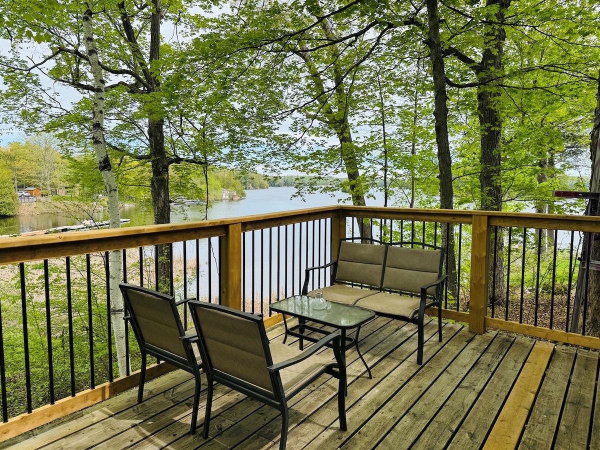 A lakeside deck is furnished with a glass-top table and a seating arrangement of four metal chairs with cushions. Lush green trees provide shade, and a serene lake is visible in the background.