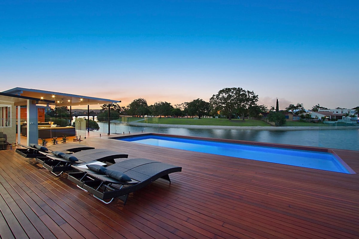 A wooden deck surrounds a sleek, heated swimming pool overlooking tranquil water and lush greenery. Modern loungers are arranged for relaxation, with ambient outdoor lighting illuminating the space as the sky transitions into dusk.