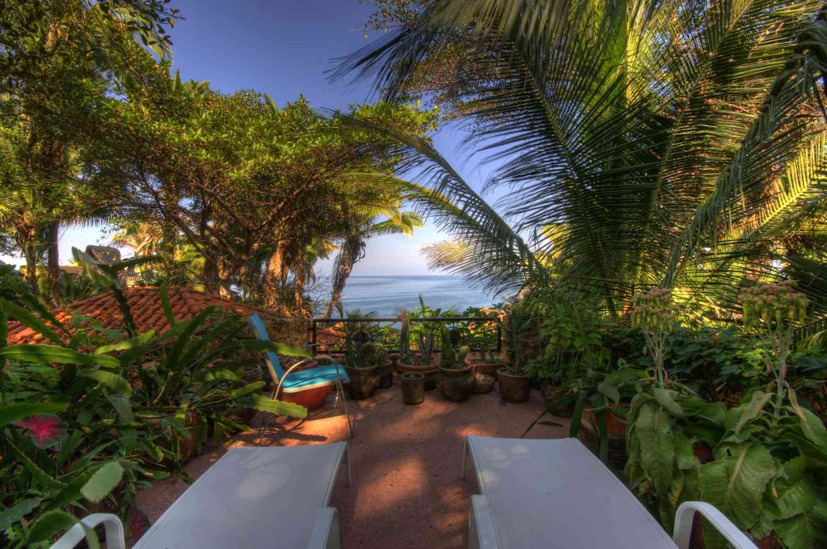 A peaceful outdoor terrace is surrounded by lush tropical vegetation, offering views of the ocean through the palm fronds. Two sun loungers are placed on the patio, inviting relaxation in a serene natural setting.