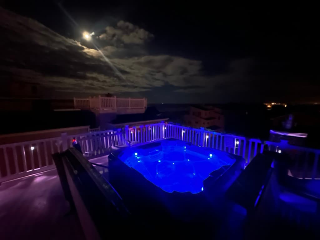 A rooftop deck features a salt-water hot tub illuminated in soft blue tones, surrounded by a white railing. The night sky showcases a full moon and scattered clouds, enhancing the tranquil ambiance. Distant lights of the town are faintly visible in the background.